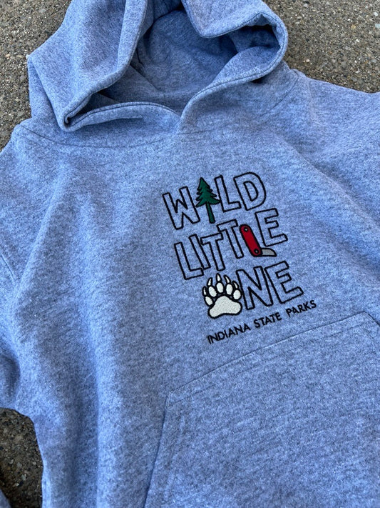 A youth grey hoodie laid flat on a gray concrete surface, featuring a front graphic that reads 'WILD LITTLE ONE' with a tree, red backpack, paw print, and 'INDIANA STATE PARKS' text.