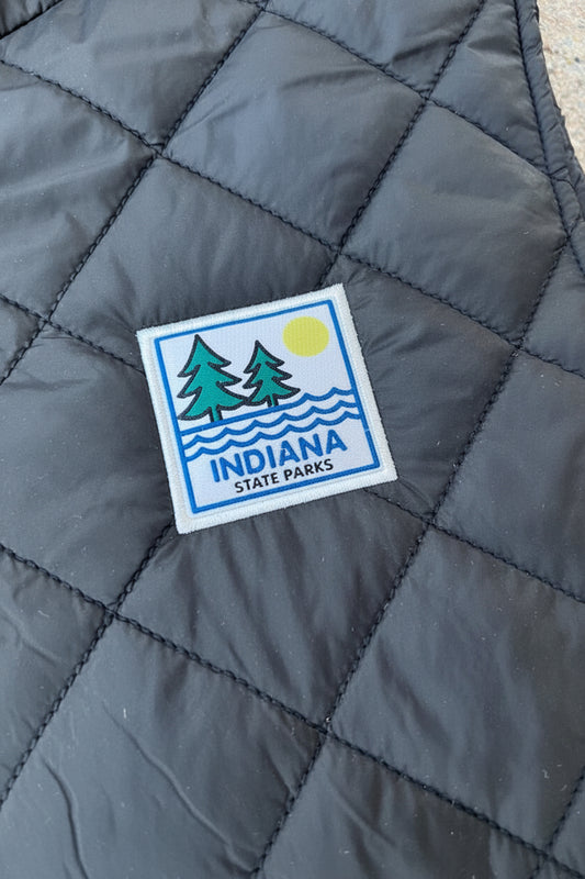 A close-up of a black quilted fabric item featuring a square patch that reads 'INDIANA STATE PARKS' with pine trees, wavy lines, and a yellow sun.