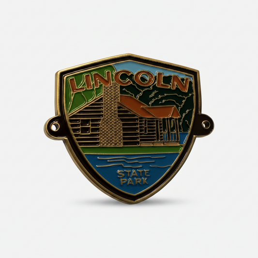 Shield shaped enamel hiking stick medallion with image of brown log cabin and blue water and 'Lincoln State Park' in text