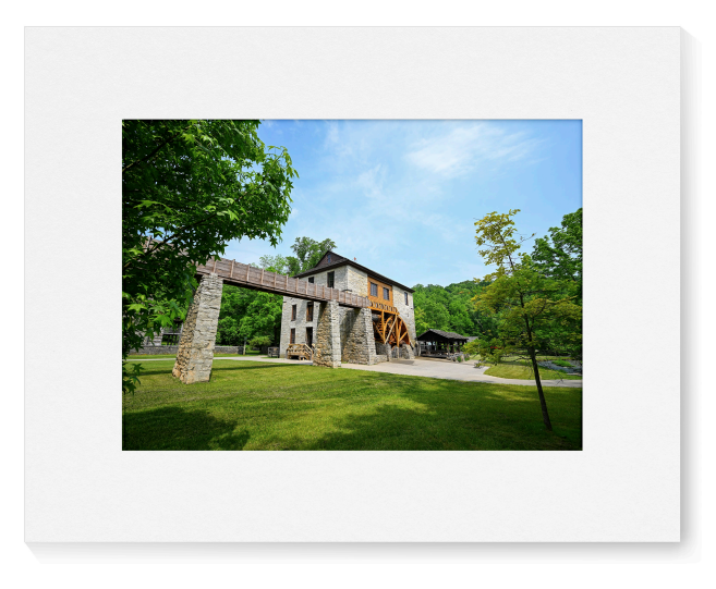 A matted art print featuring a scenic photograph of a historic stone mill with a large wooden water wheel, surrounded by green trees and grass under a blue sky.