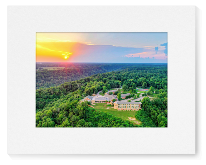 A matted art print showing an aerial landscape scene of at sunset, with a large white mat and a central rectangular print featuring dense green forest, Clifty Inn, and a glowing yellow-orange sun in the sky.