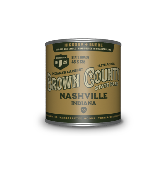 A cylindrical metal tin with Brown County State Park label, Hickory + Suede candle scent, featuring a tan and dark green label, set against a plain white background.