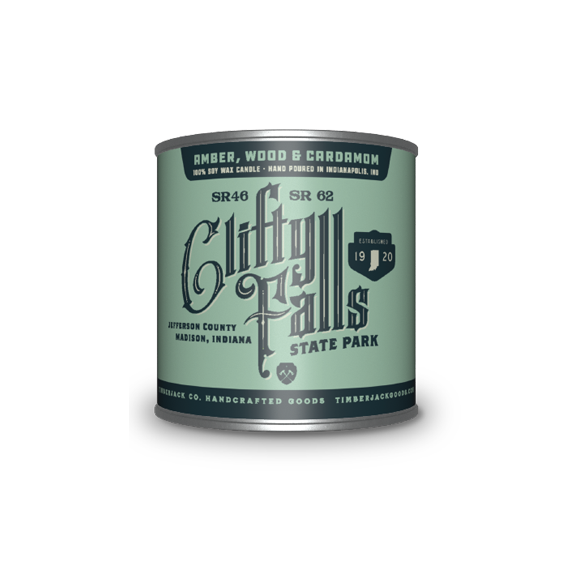 A cylindrical metal tin with Clifty Falls State Park label, Amber, Wood, and Cardamom candle scent, featuring a light green and dark green label, set against a plain white background.
