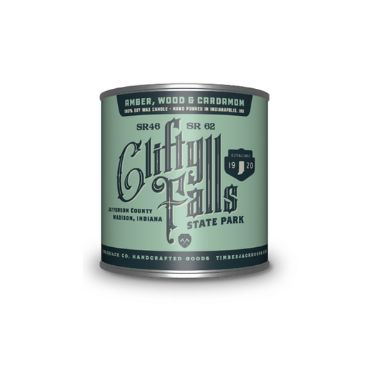 A cylindrical metal tin with Clifty Falls State Park label, Amber, Wood, and Cardamom candle scent, featuring a light green and dark green label, set against a plain white background.