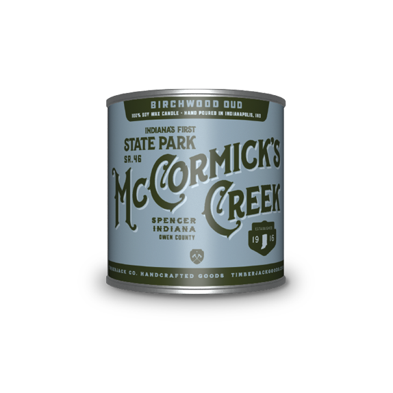 A cylindrical metal tin with McCormick's Creek State Park label, Birchwood Oud candle scent, featuring a blue and dark green label, set against a plain white background.