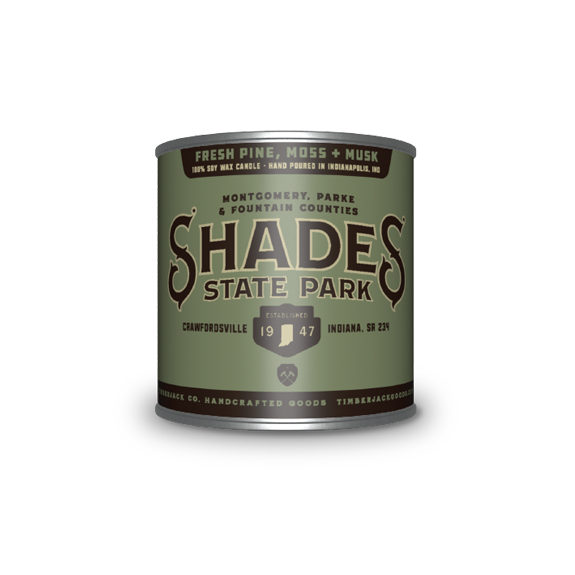 A cylindrical metal tin with Shades State Park label, Fresh Pine, Moss, and Musk candle scent, featuring a green and dark green label, set against a plain white background.
