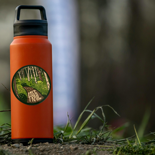 Orange water bottle with a forest design sticker on grass with a blurred forest background