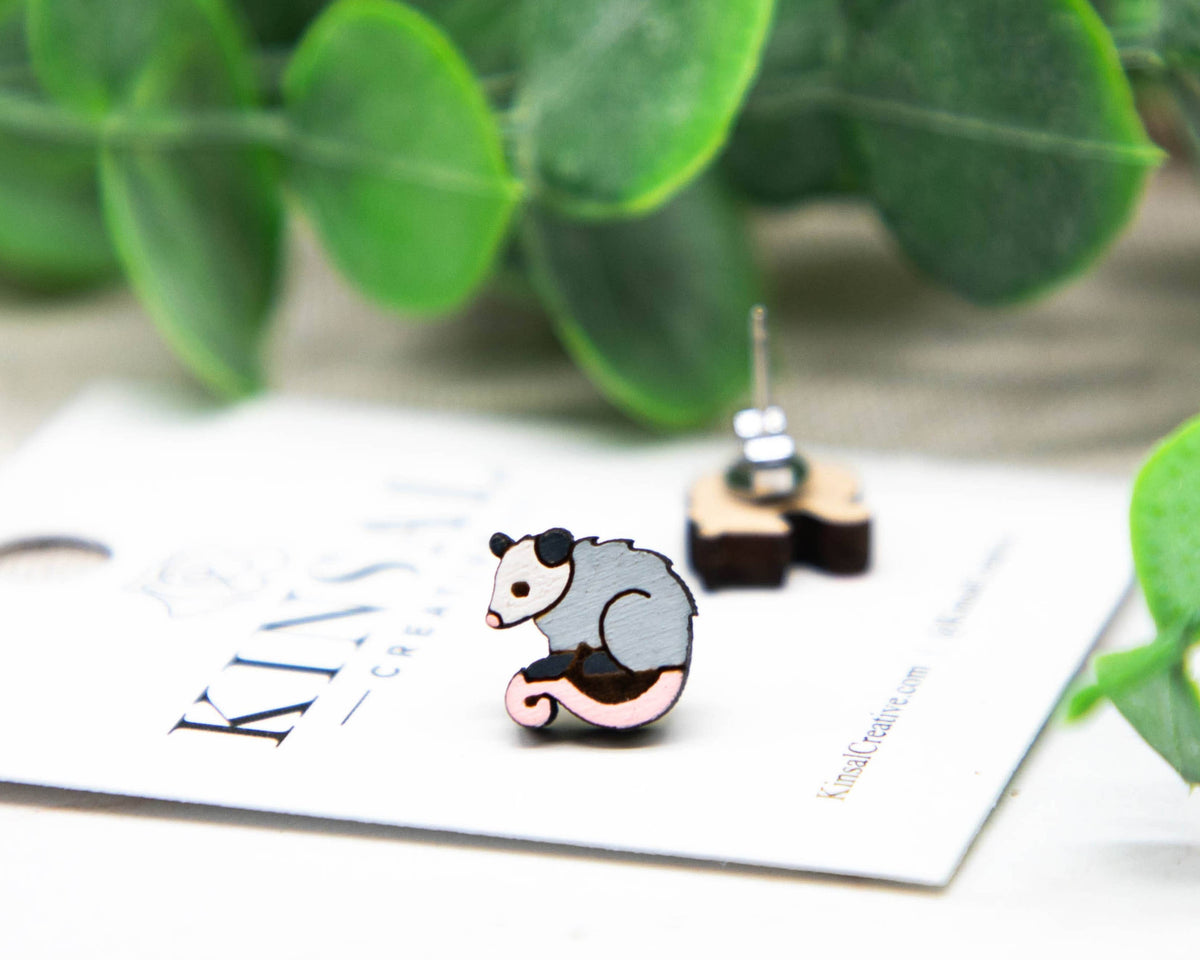 Opossum-shaped earrings on a card with green leaves in the background
