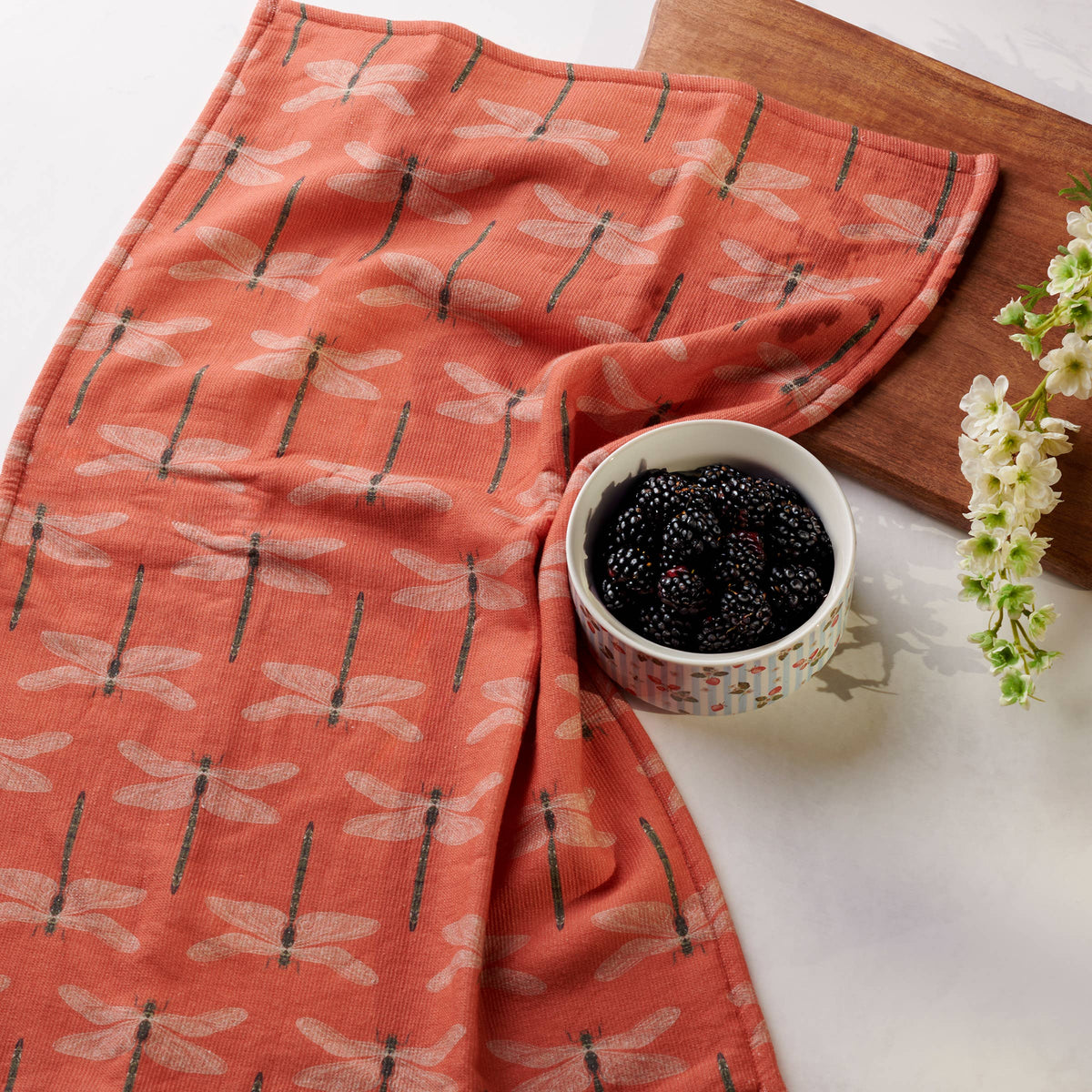 Coral-colored towel with dragonfly pattern draped over a bowl of blackberries on a white surface.