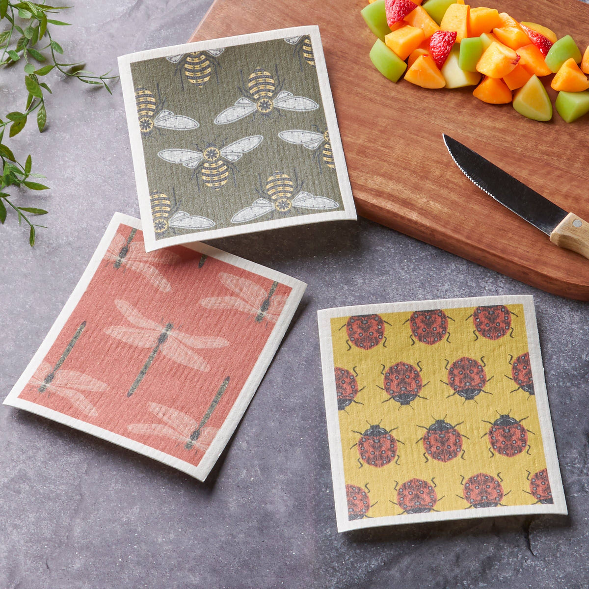 Three patterned dishcloths on a gray surface with a cutting board and fruit in the background.