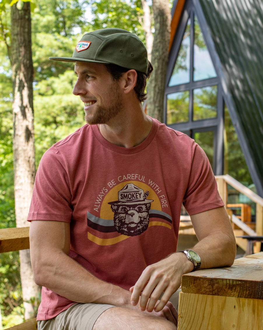 Man wearing a red t-shirt with a graphic design, sitting outdoors near a wooden structure.